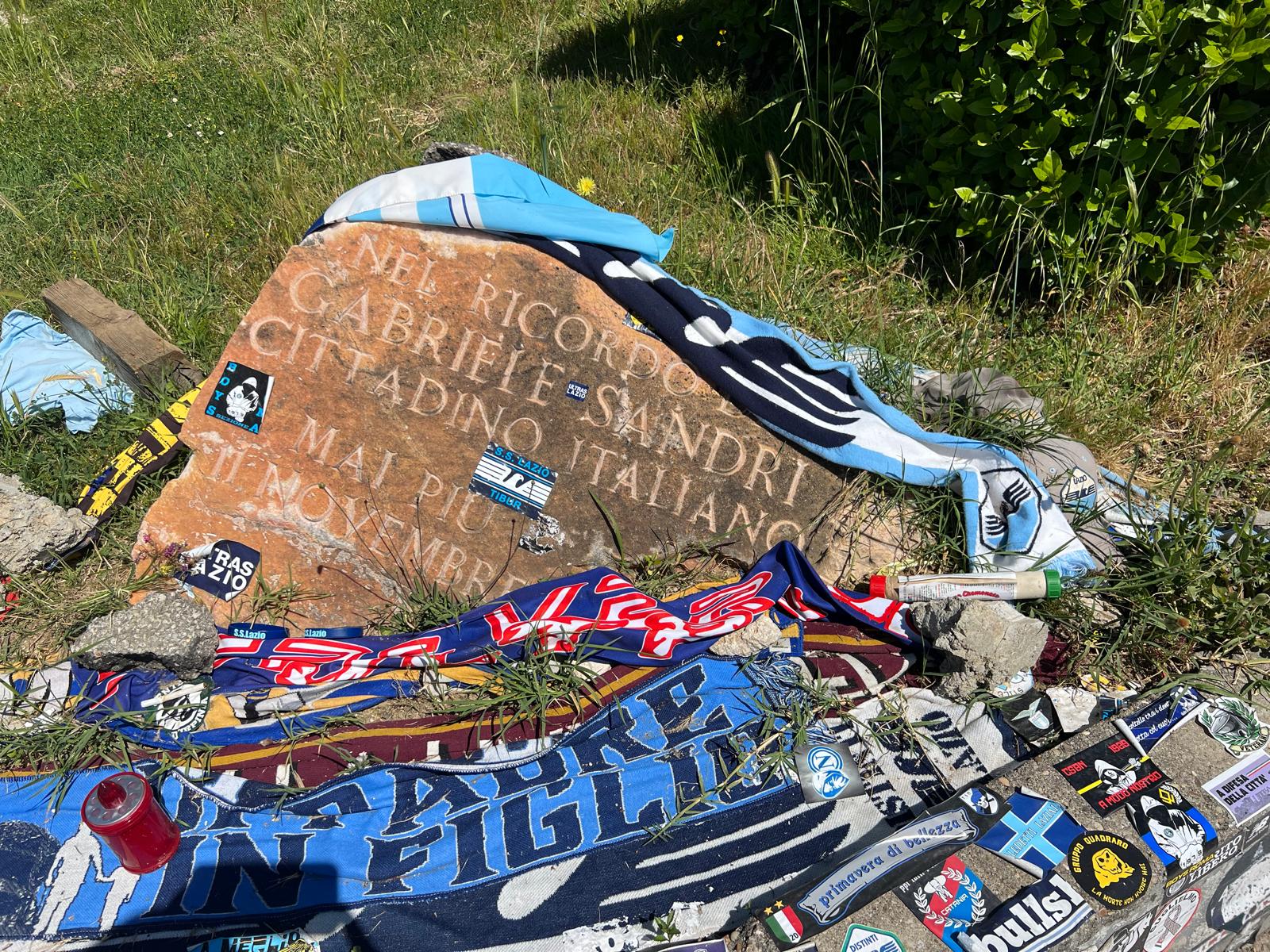 Memorial stone for Gabriele Sandri covered in football stickers and scarves from various teams, left as tributes by visiting supporters.