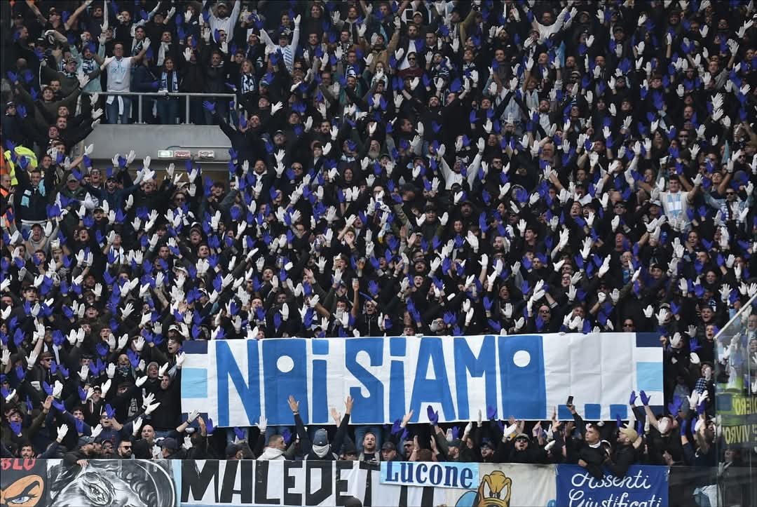 Lazio fans at the Atalanta match wearing white and blue gloves, passionately singing and waving their arms in unison from the away section.