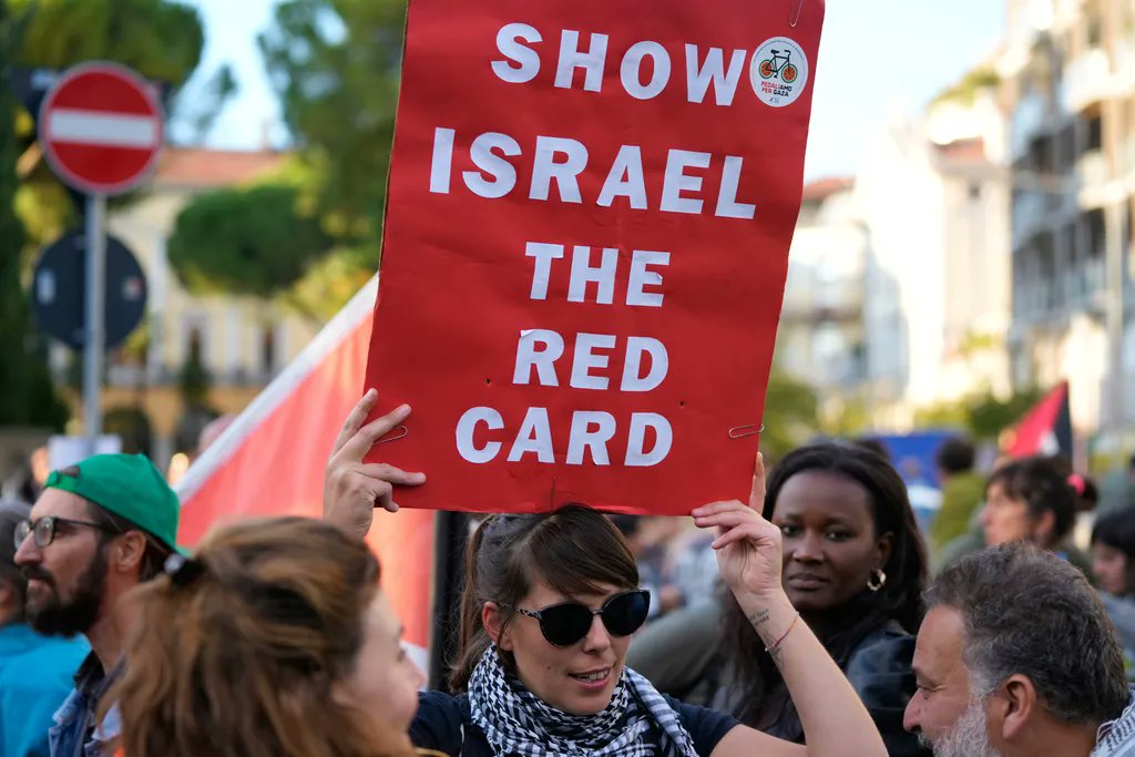 A woman holding a banner that reads "Show Israel the Red Card" during a protest or demonstration.