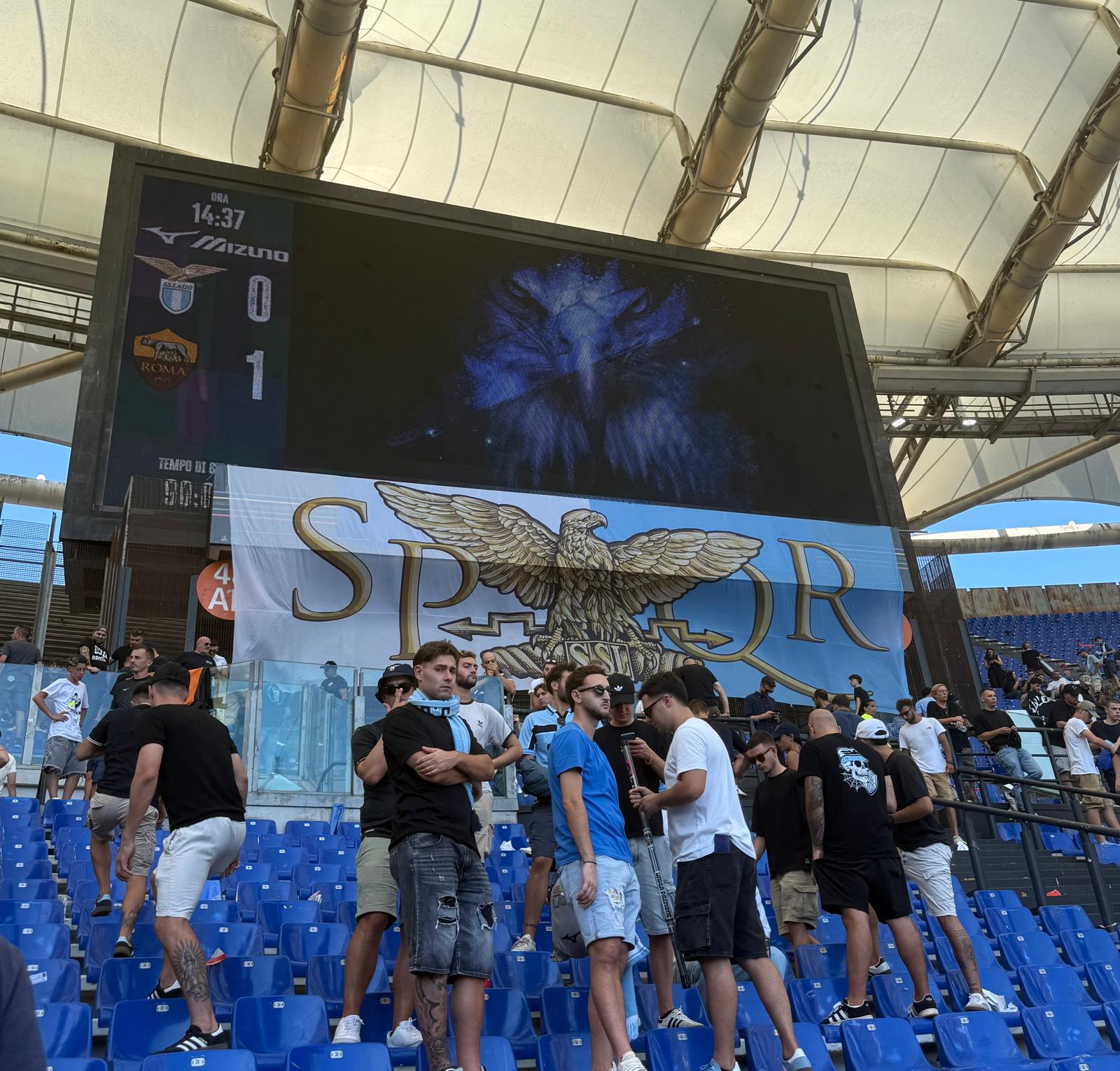 Scoreboard showing Lazio 0–1 Roma after the Derby della Capitale, with a Lazio flag displayed beneath it.