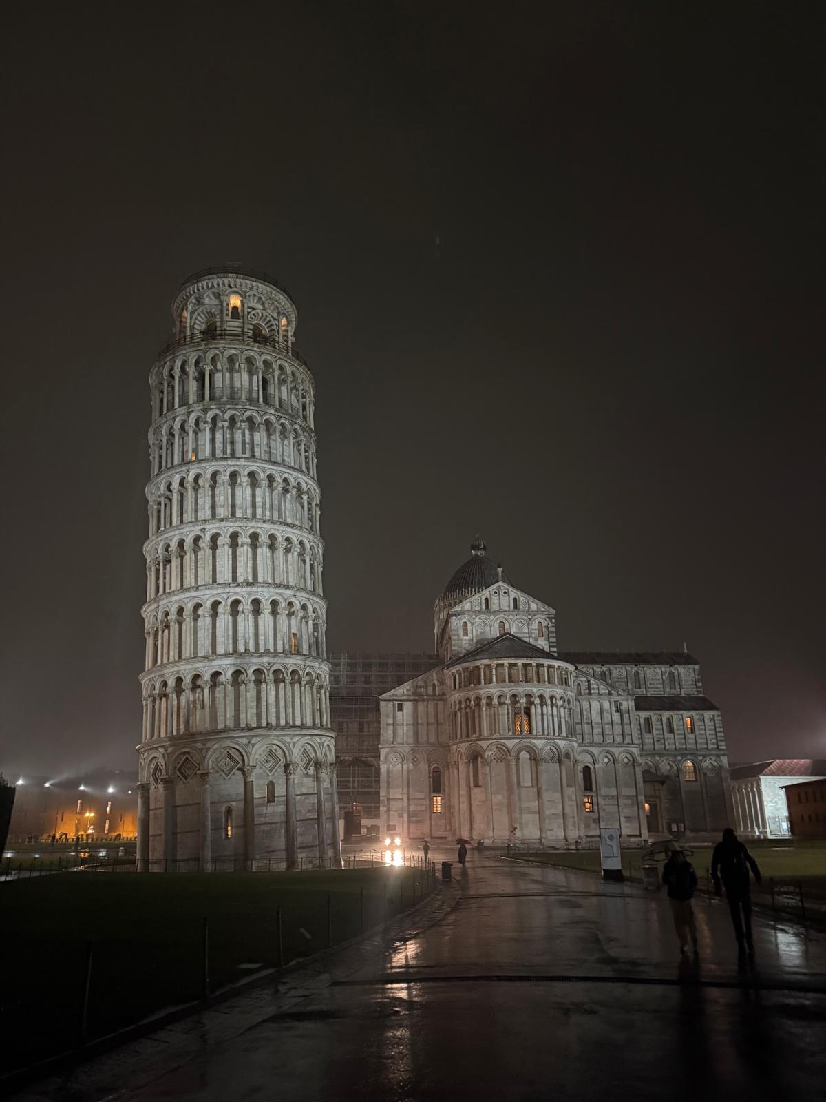 Leaning Tower of Pisa illuminated at night, glowing against a dark sky. Rain is falling and visible in the light, creating reflections on the wet ground. The tower leans clearly to the right, with soft lighting highlighting its arches and columns. Few or no people are visible, giving the scene a calm, atmospheric feel.