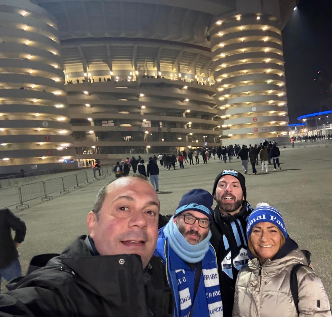Scott, Simone, Janina and Luca standing together outside the San Siro before the match, smiling in front of the stadium’s iconic red towers and spiral ramps on a bright matchday.