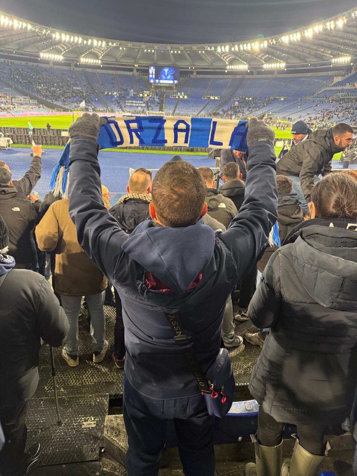 Lazio supporter in the Curva Nord holding a sky-blue and white scarf aloft among fellow fans, with banners and stadium lights visible during a match at the Stadio Olimpico.
