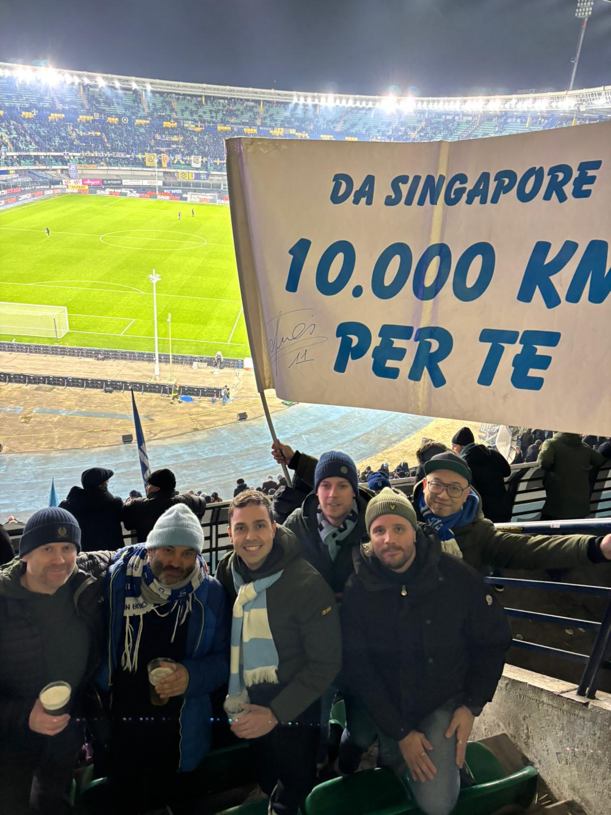 A group of S.S. Lazio supporters standing together in the stands, proudly holding up Nigel’s flag among blue-and-white scarves and banners.