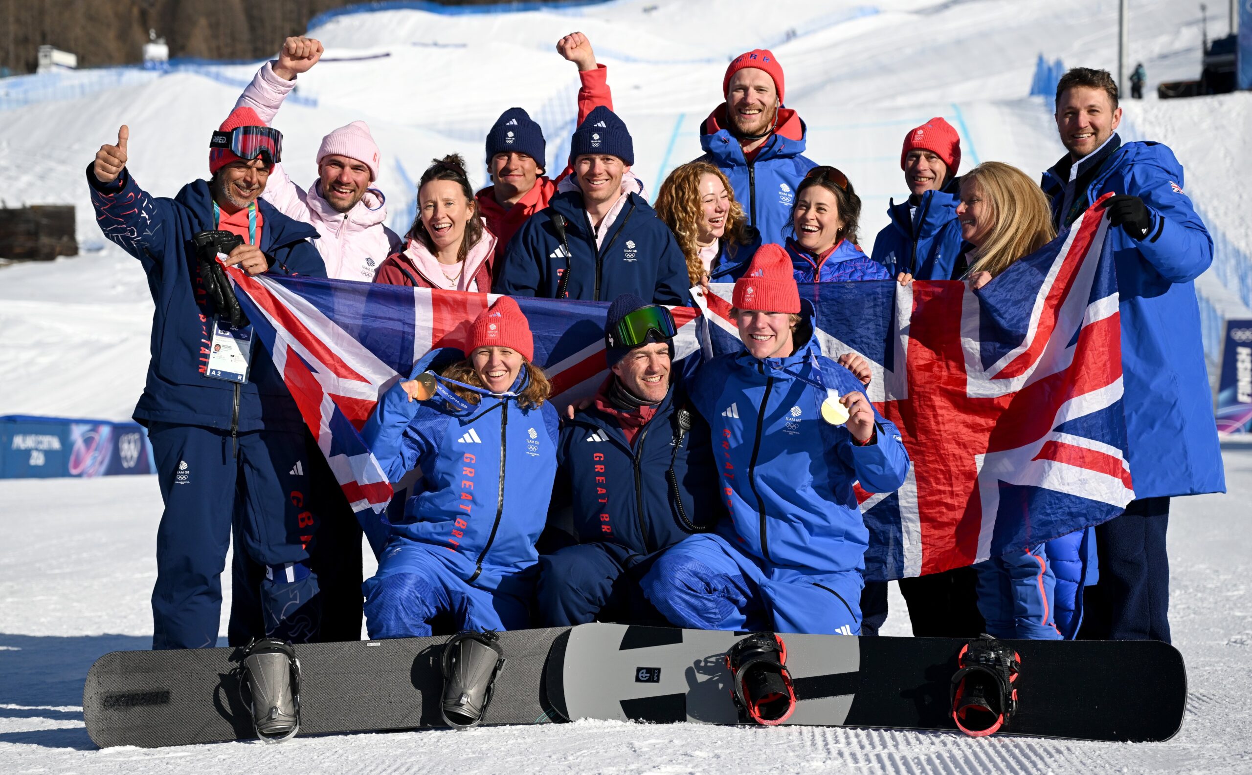 Team GB athletes standing together at the Milan-Cortina 2026 Winter Olympics, proudly wearing their gold, silver and bronze medals, draped in Union Jack flags and smiling during the medal ceremony celebration.
