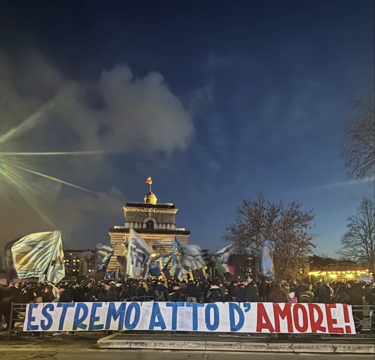 A large crowd of football supporters gathered at dusk in front of a historic stone building with a small dome and Italian flag on top. Fans wave blue and white flags while holding a long banner reading “ESTREMO ATTO D’AMORE!” in bold blue and red letters. The sky is darkening with clouds overhead, and bright lights illuminate the scene from the left.