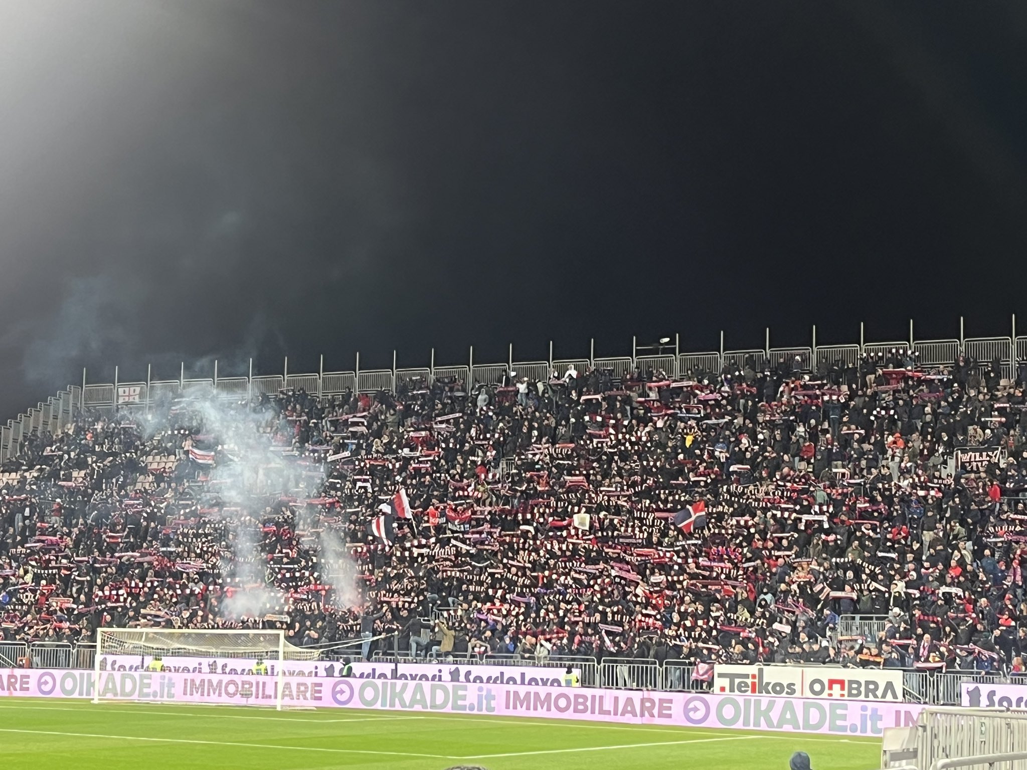 Home supporters of Cagliari packed tightly behind the goal during Cagliari vs Lazio at the Unipol Domus, flags raised and scarves aloft, creating a wall of red and blue as the teams attack the near end — photo taken by Paul Doney.