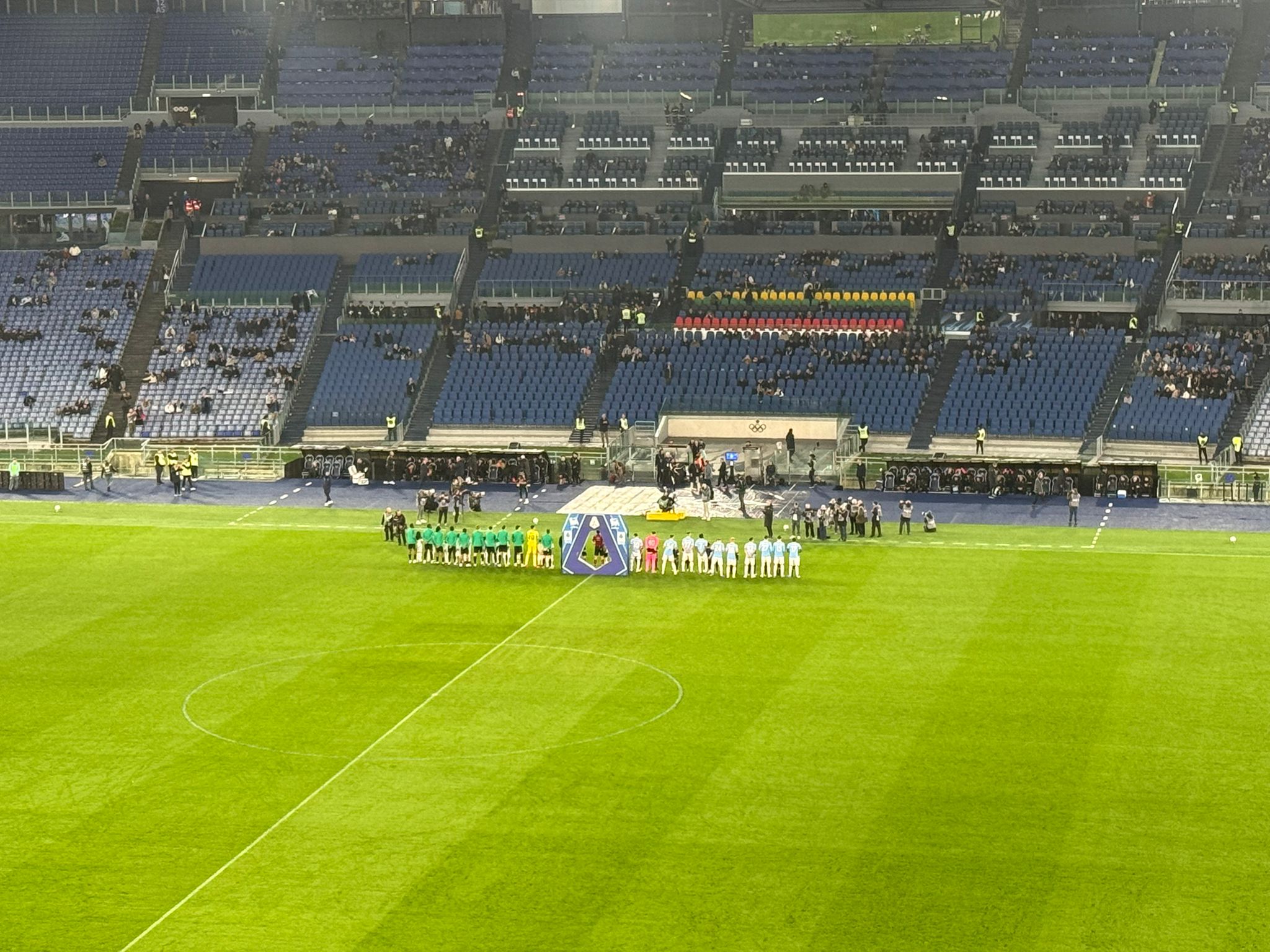 Lazio and Sassuolo players stand lined up before kick-off at the Stadio Olimpico, with large sections of empty seats visible around the stadium.