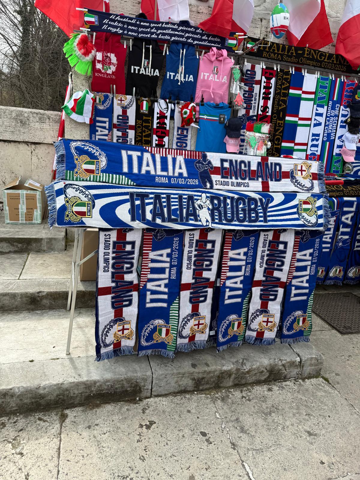 Matchday merchandise stall outside the Stadio Olimpico in Rome before Italy vs England in the Six Nations, displaying rugby scarves, hats and flags including a blue and white half-and-half scarf combining Italy and England colours.