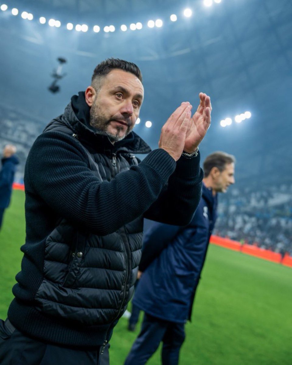 De Zerbi applauds on the touchline during a match, wearing a black padded jacket, with stadium lights glowing overhead and players and staff blurred in the background.