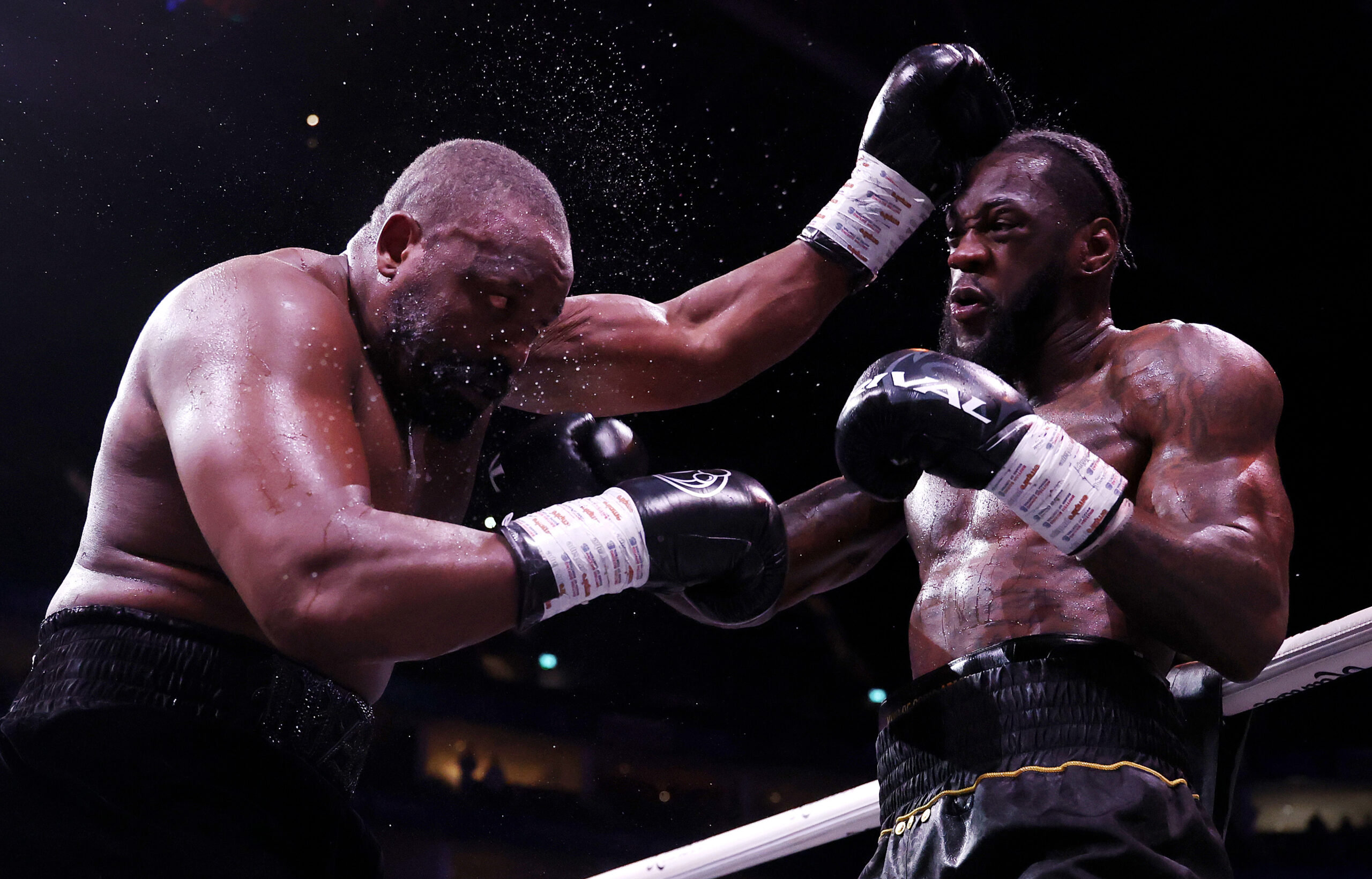 Deontay Wilder lands a powerful right hand on Derek Chisora during a heavyweight boxing match, with sweat spraying into the air as Chisora absorbs the punch near the ropes under bright arena lights.